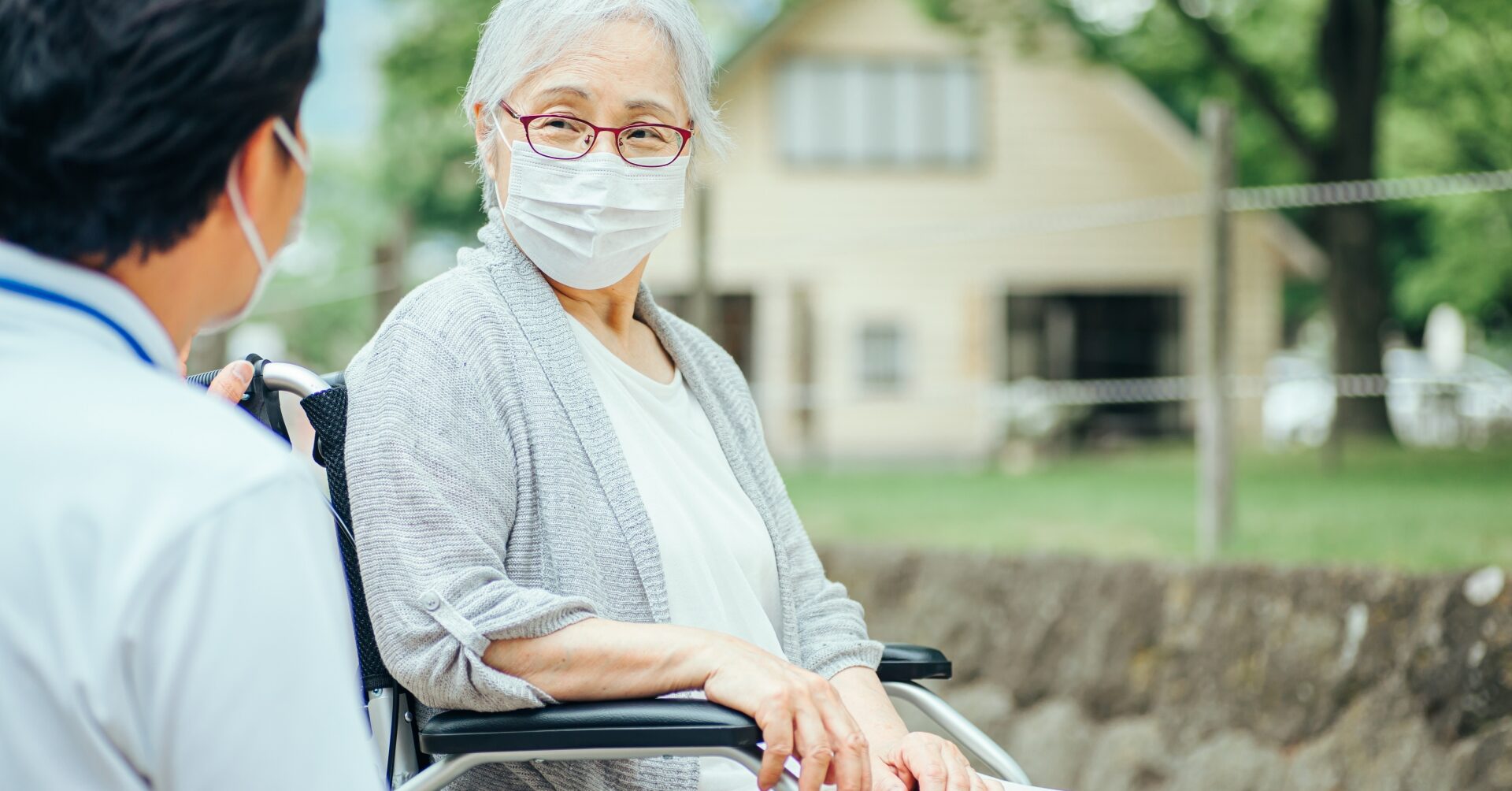 晴れた日の病院の庭で、車椅子に座る高齢の女性が、白衣の医師を穏やかな表情で見つめています。