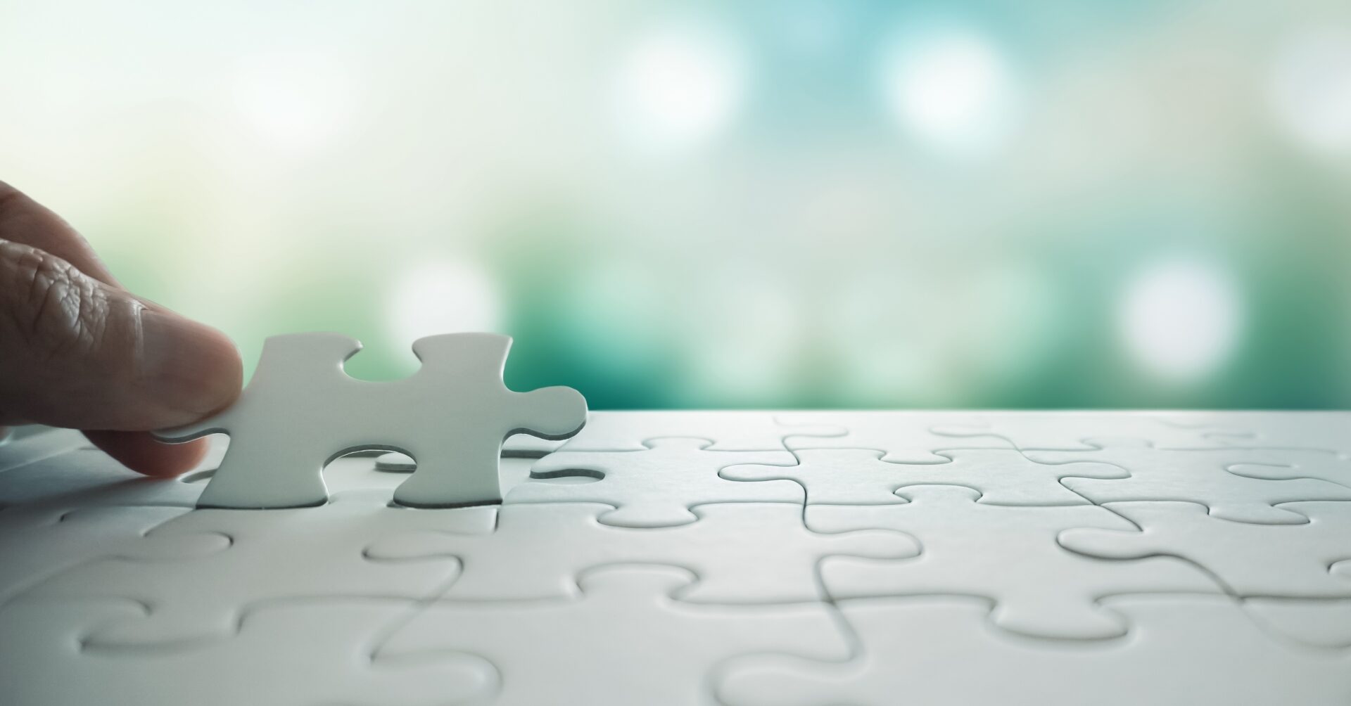 白いパズルのピースを手で最後にはめ込もうとしているクローズアップ写真。背景は柔らかなグリーン～ブルーのぼかし光で、希望や解決への一歩を象徴するイメージ。