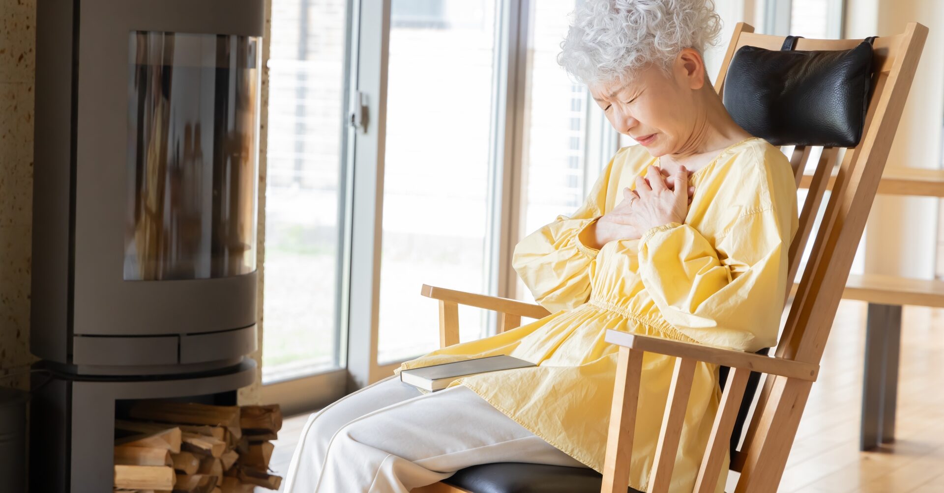 ロッキングチェアに座り、胸元を押さえて苦痛に顔をしかめる高齢の女性。慢性疼痛を抱える状況を表す。